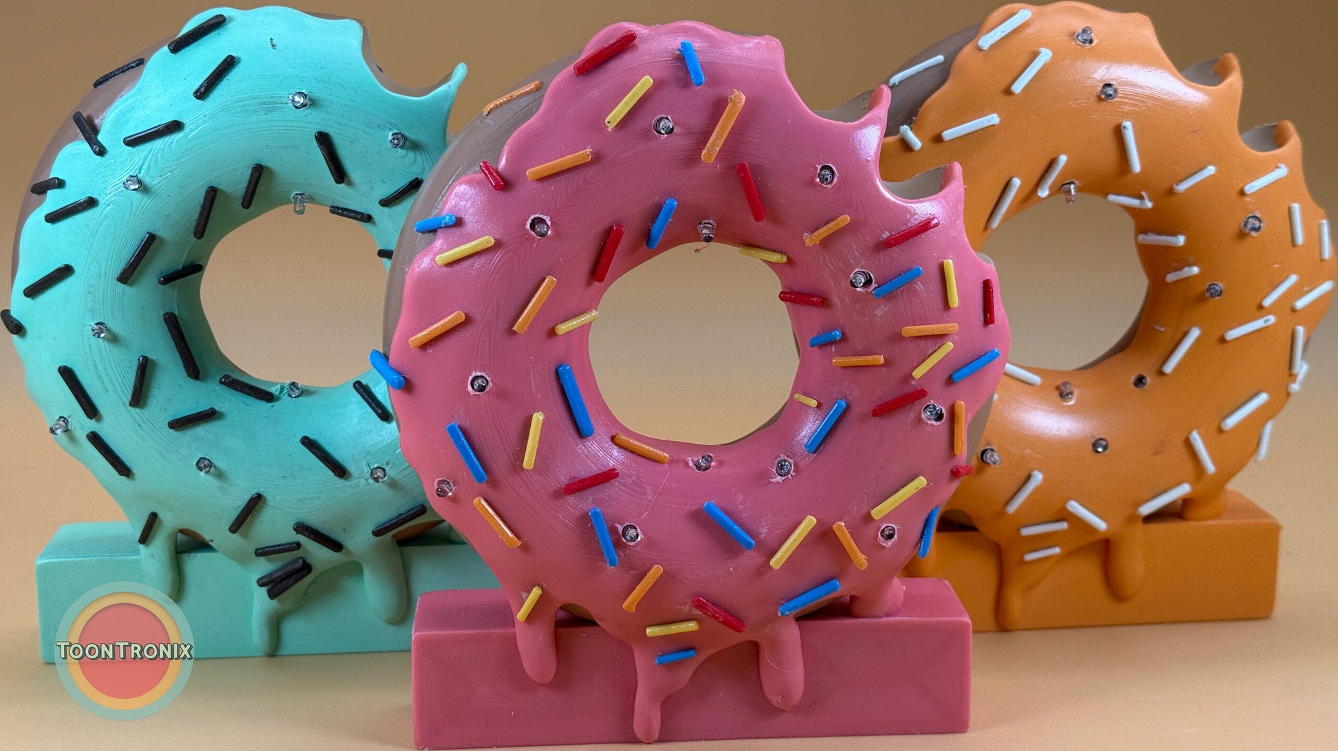 Colorful 6" or 9" donut-shaped desk lamps with sprinkles in pink, teal, and orange designs on a beige surface