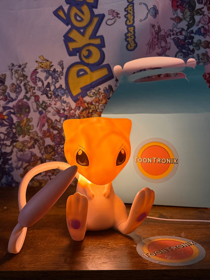 7.5” Mew Desk Lamp figure with movable tail and USB cord glowing on wooden surface with Pokemon backdrop and Toontronix packaging